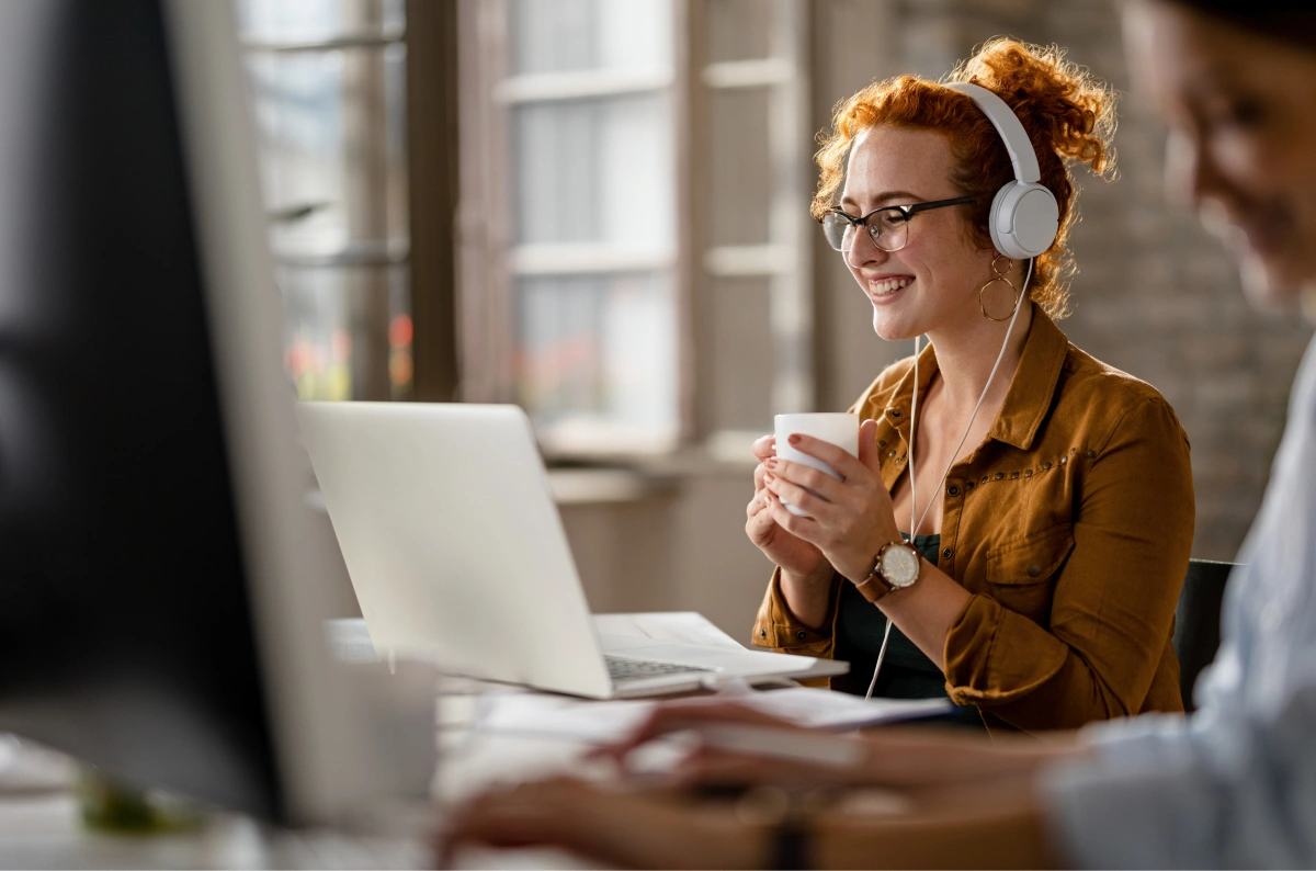 happy-female-entrepreneur-wearing-headphones-while-surfing-net-laptop-drinking-coffee-office 1