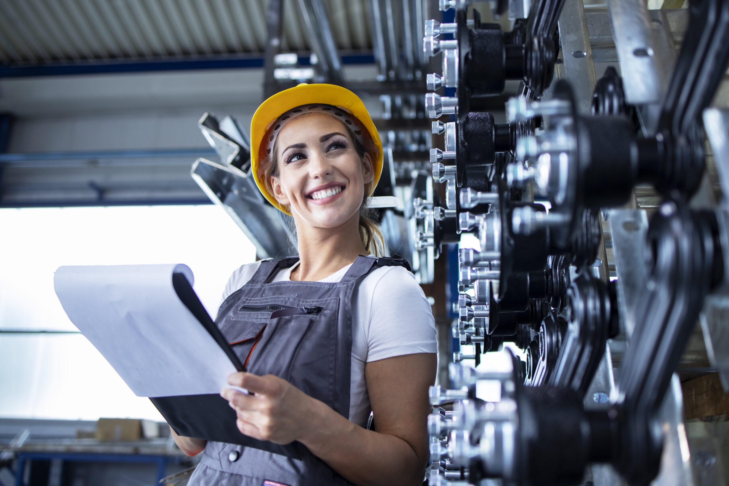 Portrait of female industrial employee in working uniform and hardhat checking production in factory.