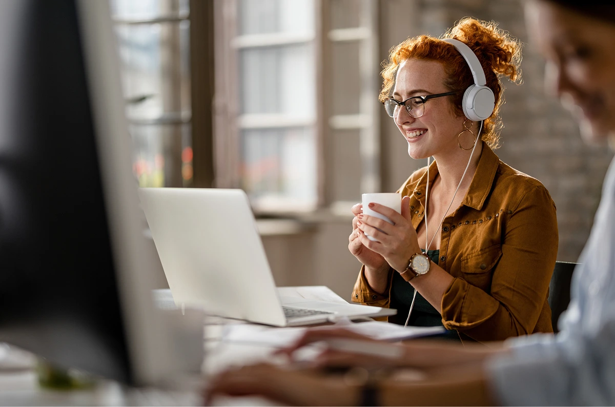 happy-female-entrepreneur-wearing-headphones-while-surfing-net-laptop-drinking-coffee-office 1 (1)