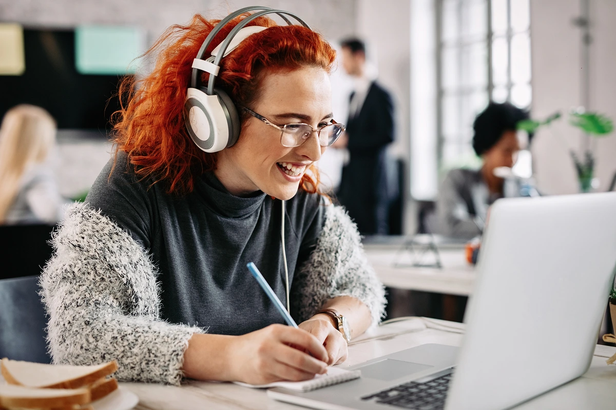 cheerful-female-entrepreneur-working-computer-taking-notes-her-notepad-while-listening-music-headphones-office-there-are-people-background 1