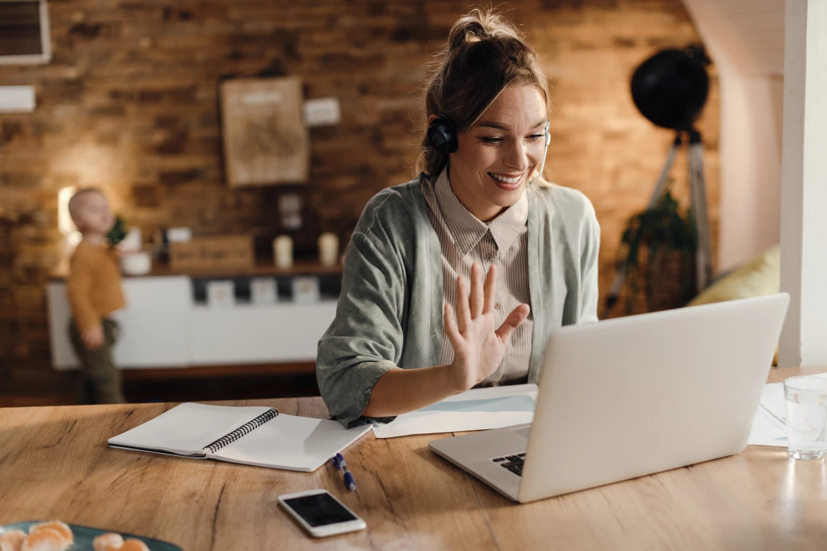 happy-female-entrepreneur-waving-video-call-laptop-home 1